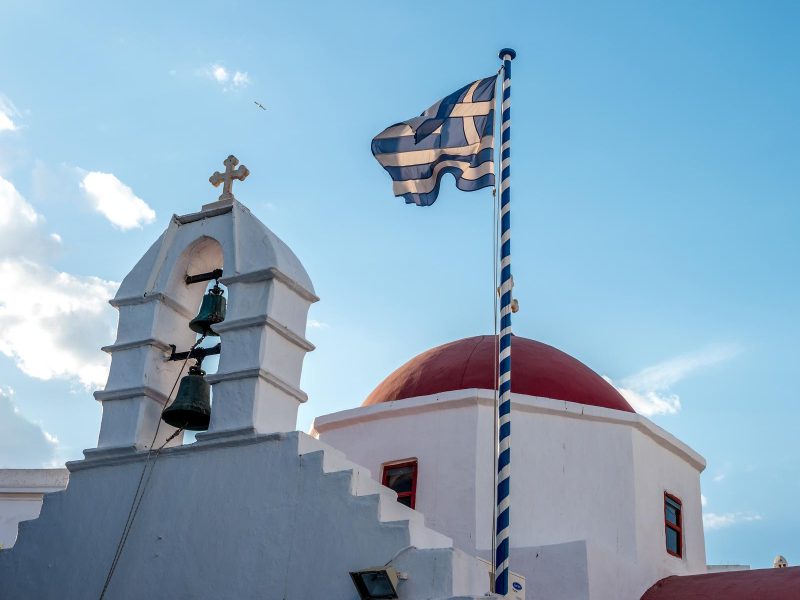 Agia Kyriaki Orthodox church with red dome and Greek flag waving against blue sky on the Mykonos island, Greece